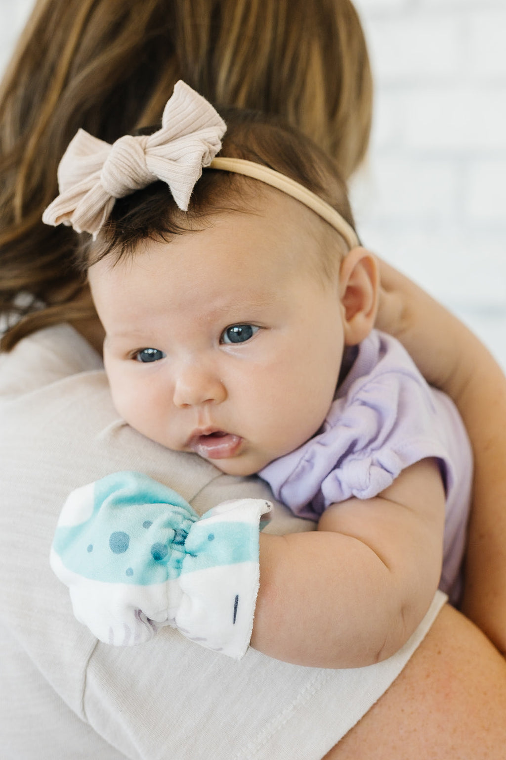 Baby girl with a tan bow, purple onesie, and dinosaur baby mittens being held by an individual with shoulder length brown hair and a white short sleeve shirt