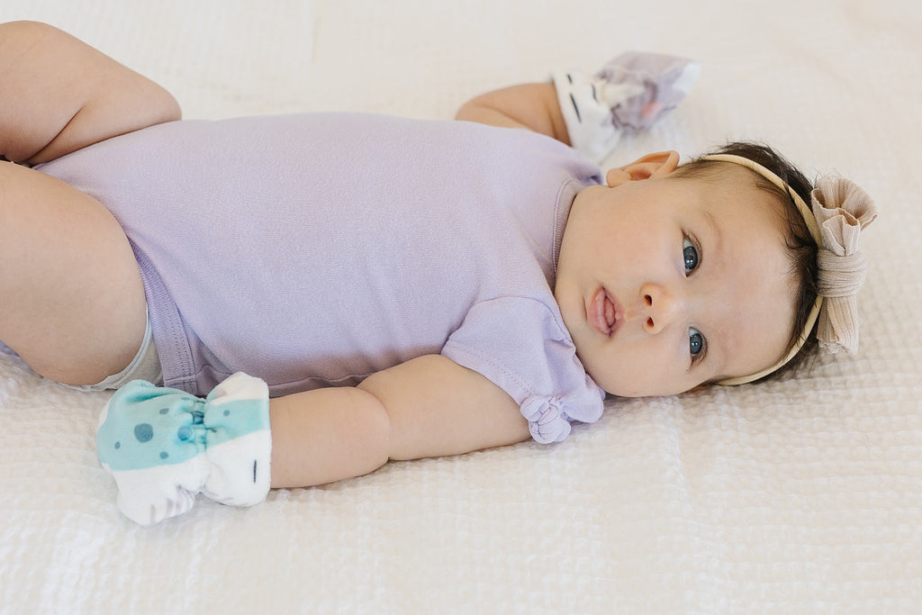Baby girl laying on a white blanket in a purple onesie wearing baby mittens with dinosaur characters